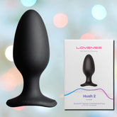 A black, silicone Lovense Hush 2 Vibrating Butt Plug - Large is placed next to its packaging. The white box prominently displays the "Lovense" brand and emphasizes its Bluetooth connectivity, remote-control capabilities, and user-friendly magnetic charging port. Pastel tones softly blur the background.